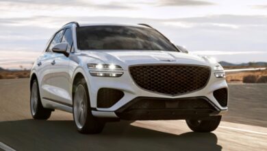 A white 2023 Genesis GV70 small luxury SUV is driving on the road.