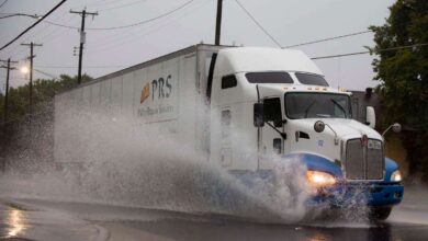 A semi truck driving through standing water and slowing down, trees visible in the background.