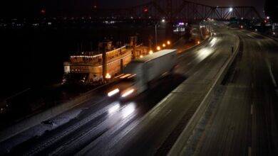 A semi truck driver flashing their high beam headlights on the interstate highway at night, a city skyline visible in the backgound.