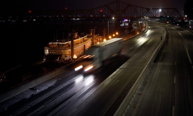 A semi truck driver flashing their high beam headlights on the interstate highway at night, a city skyline visible in the backgound.