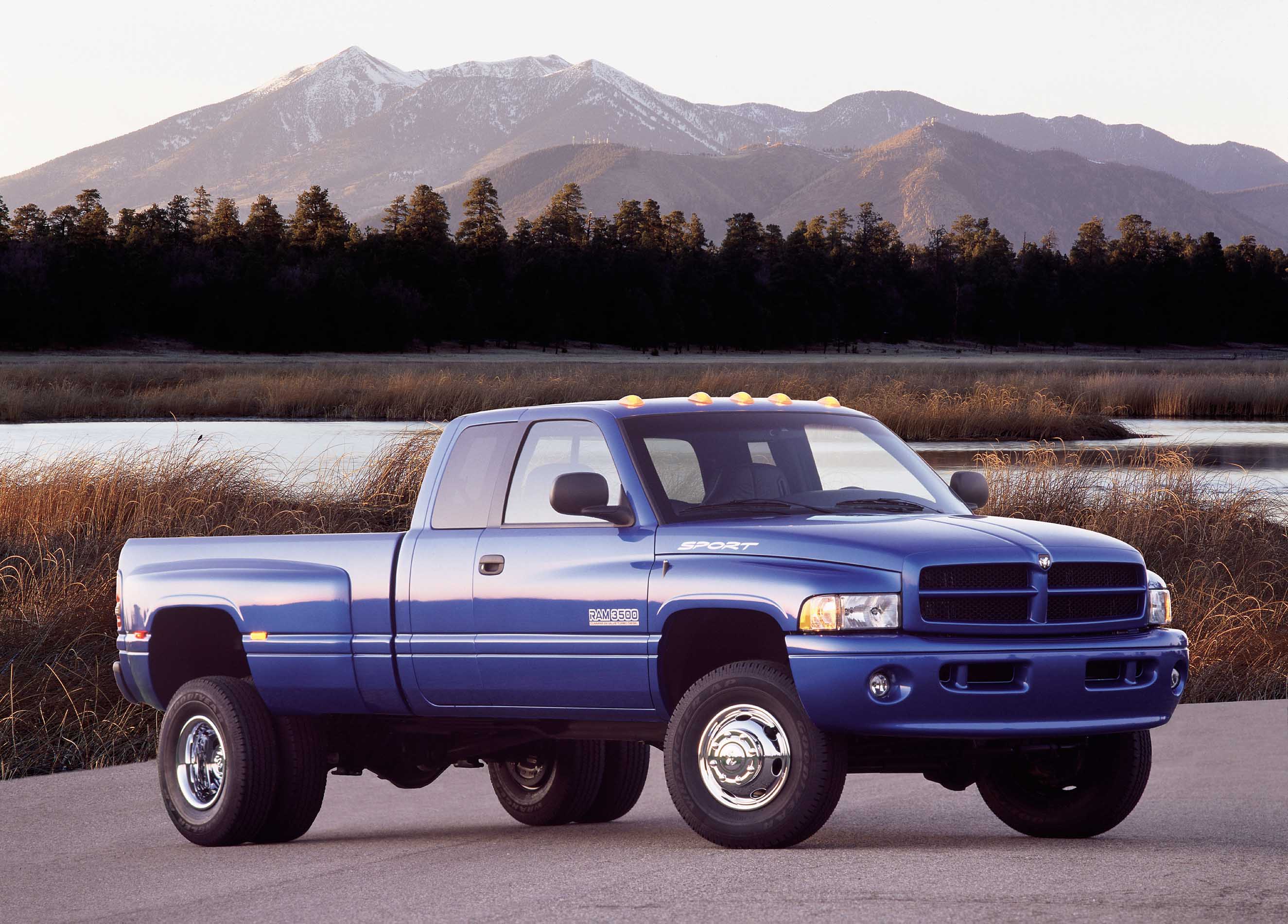 A blue 2001 Dodge Ram pickup truck with a second generation Cummins engine is parked by a lake with mountains in the background.
