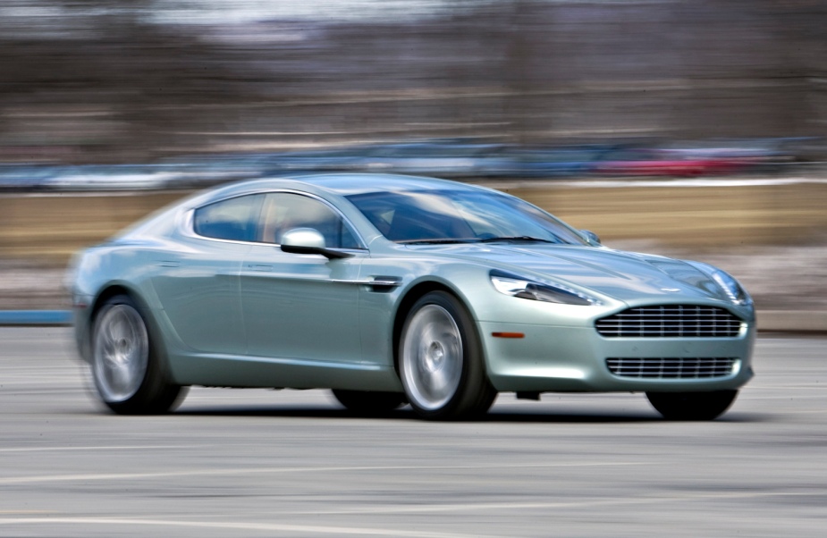Water green Aston Maritn Rapide S sedan with four-door camera blur, row of parked cars visible in the background.