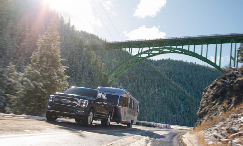 A black Ford F-150 PowerBoost hybrid pickup shows off its truck of the year powertrain by pulling a heavy camper trailer up a steep hill, a bridge visible in the background.