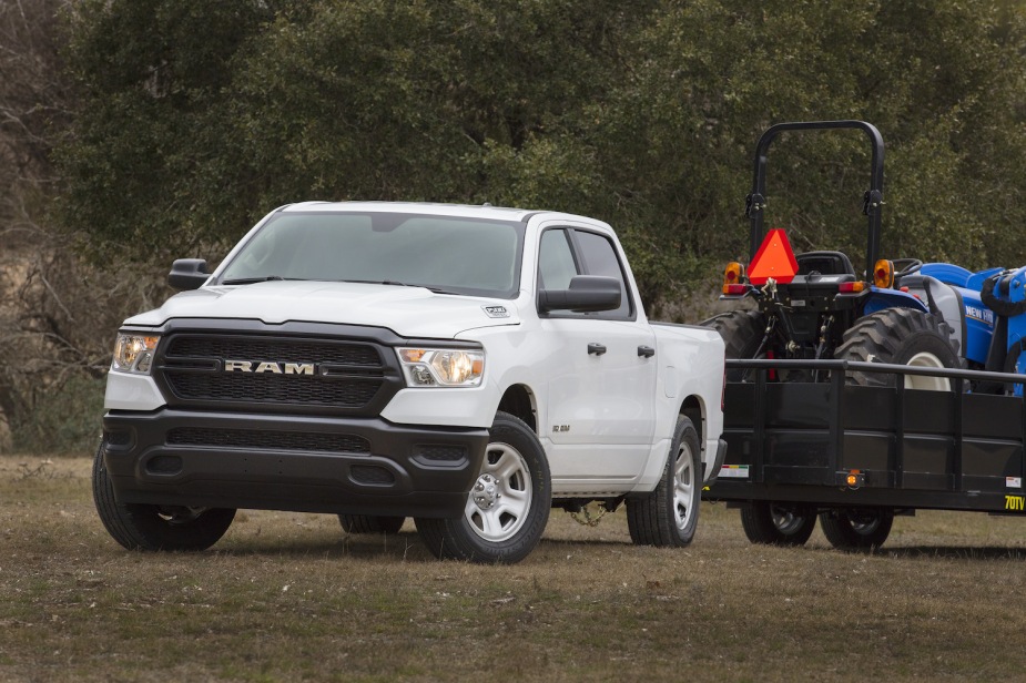 A white Ram 1500 Tradesman pickup truck, parked on a grassy field and attached to a trailer pulling a blue tractor, trees visible in the background.