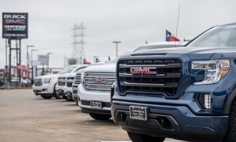 The grilles of a row of GMC and Chevrolet pickup trucks parked at a General Motors dealership, a row of buildings visible in the background.
