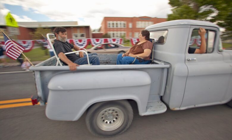 A couple rides in the bed of a primer gray stepside pickup truck during a parade.