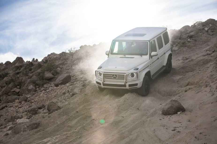 White Mercedes Benz G-Class driving down a rocky hill, blue sky in the background.