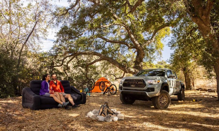 A couple sits by a campfire in the woods, their Toyota Tacoma tent visible in the background.