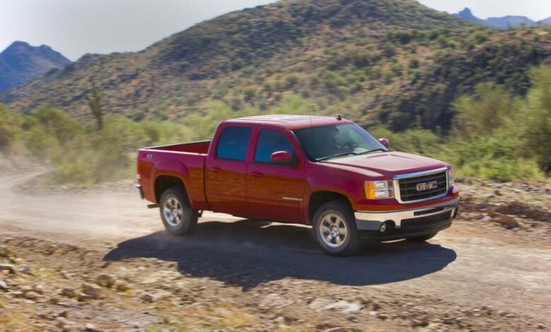 A red GMC Sierra 1500 pickup truck driving up a dirt road, mountains visible in the background.