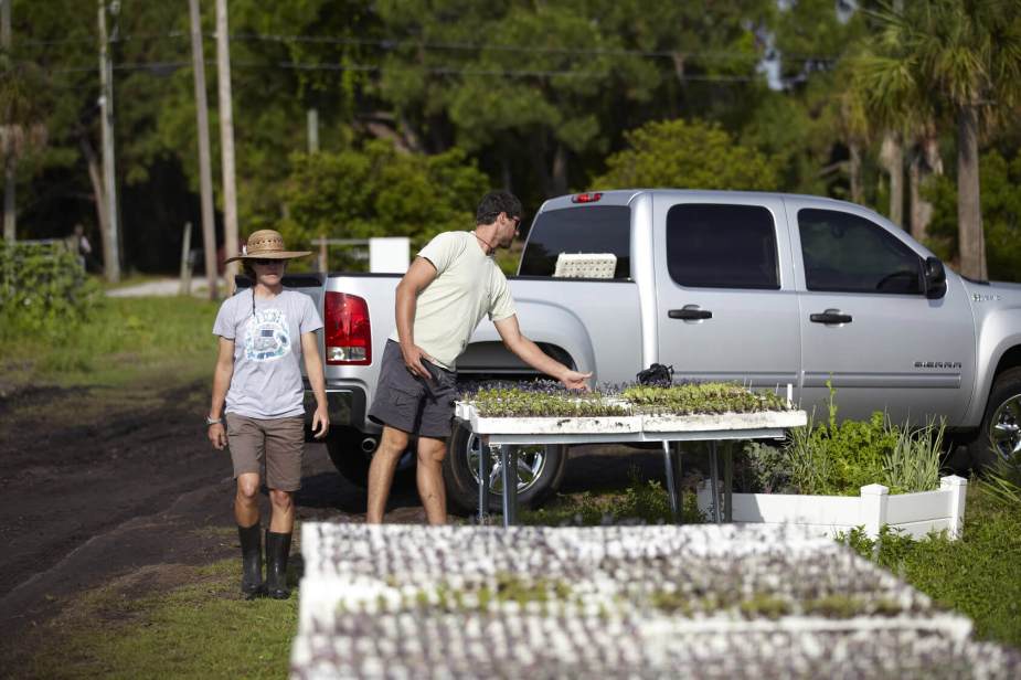 A couple stands next to trays of starter seedlings, a silver Sierra 1500 pickup truck visible behind them.