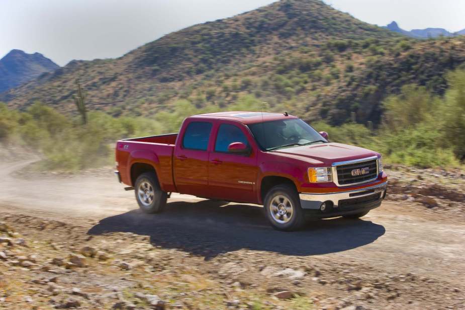 A red GMC Sierra pickup truck drives on a dirt road with trees and a mountain bottom line visible in the background.