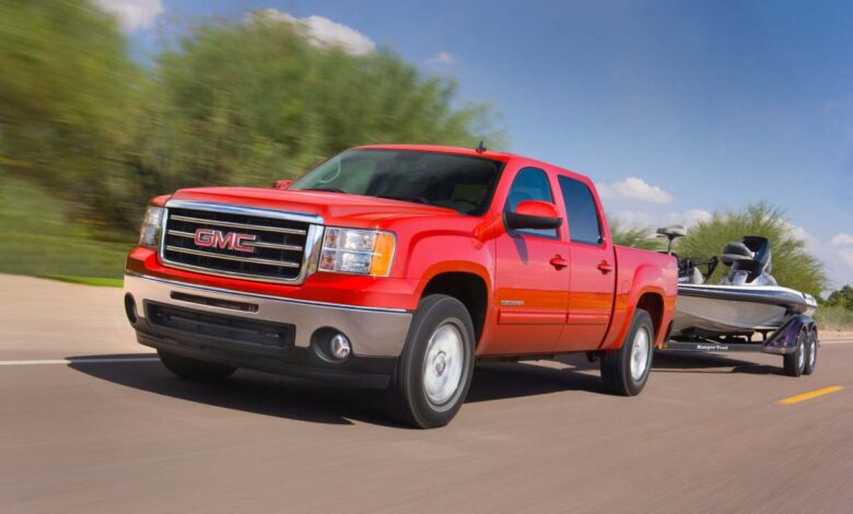 Promo photo of a red 2013 GMC Sierra 1500, a truck with relatively few engine issues, towing a boat down a rural road with trees in the background.