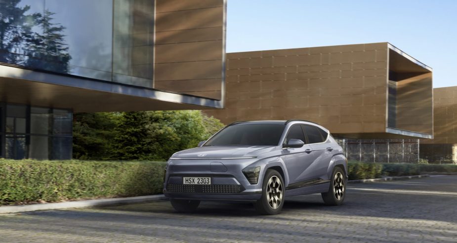 Three-quarter front view of a 2024 Hyundai Kona sitting motionless in front of a building with late day greenery and shadows. 