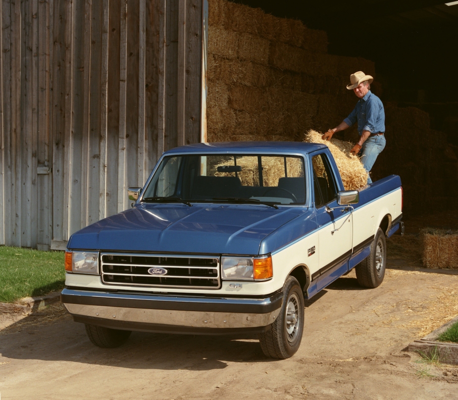 A man carries hay bales into the bed of a blue and white Ford truck.