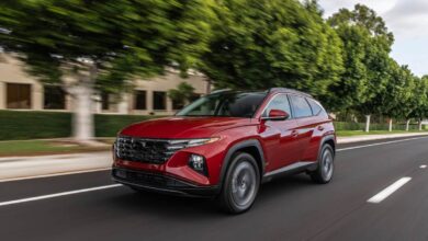 A red 2023 Hyundai Tucson SUV cruises down a road with green trees beside it. Hyundai has been cited as the year