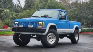 A blue 1990 Jeep Comanche Cherokee-based 4WD pickup truck in a parking lot for an auction photoshoot.