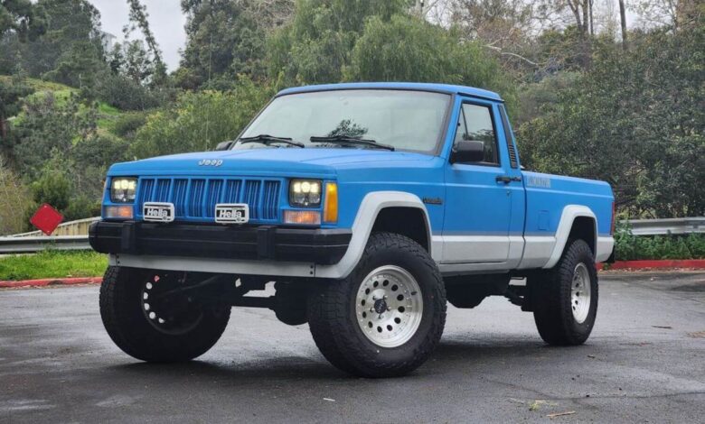 A blue 1990 Jeep Comanche Cherokee-based 4WD pickup truck in a parking lot for an auction photoshoot.